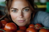 Young Caucasian woman with blue eyes and natural makeup smiling while lying among fresh red tomatoes, close-up portrait showing genuine happiness and healthy lifestyle.