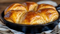 Freshly baked golden brown dinner rolls in cast iron skillet, with buttery glazed tops and soft layered texture, photographed in natural light on striped cloth.