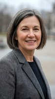 Middle-aged woman with gray hair wearing gray blazer and black shirt, smiling outdoors with blurred background for corporate or professional headshot use.