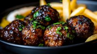 Glazed meatballs with fresh herbs and golden french fries in cast iron skillet. Comfort food dinner with crispy potato sides and parsley garnish.