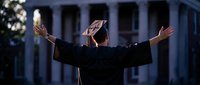 Graduate celebrating achievement with raised arms in front of academic building at dusk, wearing cap and gown for commencement ceremony success.