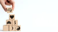 Hand placing wooden cube with brain symbol on pyramid of blocks featuring wellness icons including heart, lotus, semicolon, yin yang on white background.
