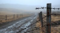 Barbed wire fence along misty rural dirt road at dawn, creating atmospheric moody landscape with rustic metal post in foreground and foggy fields beyond.