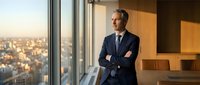 Mature male professional in dark business suit and glasses standing by floor-to-ceiling window in modern high-rise office, arms crossed, overlooking cityscape at sunset for corporate leadership and business success concepts.