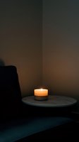 Glowing candle on round table creates warm ambient lighting in modern minimalist living room corner with dark furniture and moody atmosphere.