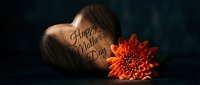 Wooden heart with engraved Mother's Day message alongside vibrant orange chrysanthemum flower on dark background for greeting cards and celebrations.