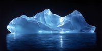 Majestic blue iceberg floating in dark ocean waters with ethereal glow, reflecting moonlight on calm sea surface. Natural arctic landscape at night creates serene atmosphere.