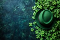 Green felt hat surrounded by fresh shamrock leaves on dark teal textured background. St Patrick's Day festive composition with copy space for holiday designs.