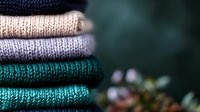 Neatly folded stack of cozy knitted sweaters in pastel and turquoise colors on dark background with soft bokeh lights and winter holiday decorations.
