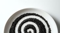 Concentric circles of black sesame seeds arranged in spiral pattern on white ceramic plate, suitable for food styling and culinary presentations.