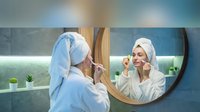 Young Caucasian woman in white bathrobe and towel turban applying cucumber slices to face while looking in bathroom mirror, skincare routine.
