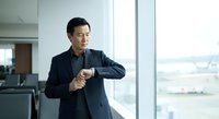 Asian male professional in dark business suit checking wristwatch in modern office with floor-to-ceiling windows, suitable for corporate, business, and time management concepts.