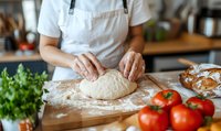 Female baker kneading fresh dough on wooden board with fresh tomatoes and herbs in kitchen. Homemade bread preparation process showing traditional baking techniques.