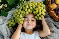 Young child with closed eyes wearing green grapes as a crown, surrounded by fresh fruit and wicker baskets on neutral fabric. Healthy eating and playful food concept.