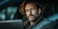 Middle-aged Caucasian man with curly hair and beard sitting in car interior, contemplative expression, automotive lifestyle portrait for business concepts.