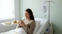 Young woman patient relaxing in hospital bed using smartphone while receiving IV treatment in modern medical facility for recovery and communication.
