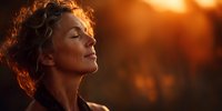 Peaceful middle-aged woman with curly hair meditating outdoors during golden hour sunset, eyes closed in serene contemplation for wellness and mindfulness.