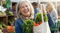 Happy senior Caucasian woman holding reusable shopping bag with fresh vegetables at farmers market, smiling while buying organic produce for healthy lifestyle.