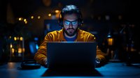 Caucasian male programmer in yellow hoodie working late at night on laptop, wearing headphones and glasses, illuminated by screen in dark office space.