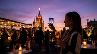 Woman with long dark hair photographing illuminated castle at twilight during evening celebration with crowds and golden lights surrounding the venue.