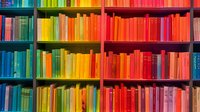 Colorful rainbow bookshelf arrangement with books organized by color spectrum from blue to red creating vibrant gradient pattern in library or bookstore interior.