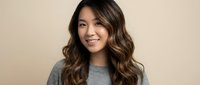 Young Asian woman with long wavy brown hair wearing casual gray sweater smiling at camera against neutral beige background for lifestyle portrait.