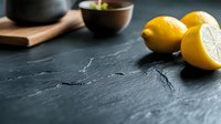 Fresh yellow lemons on dark slate surface with wooden spoon and bowl in background for cooking and food preparation in modern kitchen setting.