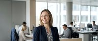 Middle-aged woman with brown hair, Caucasian ethnicity, wearing dark blazer and patterned blouse, smiling confidently in modern office environment with colleagues working at desks in background, suitable for corporate, business leadership and workplace themes.