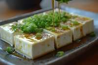 Fresh silken tofu blocks drizzled with soy sauce and sesame oil, garnished with chopped green onions on ceramic plate for healthy Asian cuisine and vegetarian cooking.