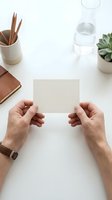 Male hands holding blank beige card on white desk with notebook, glass of water and succulent plant. Mockup template for business cards, invitations.
