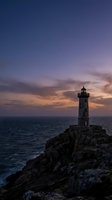 Dramatic lighthouse standing on rocky cliff overlooking ocean at sunset with purple and orange sky creating moody seascape for maritime navigation concepts.