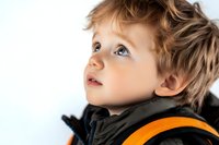 Young boy with blue eyes and tousled light brown hair looking up with wonder and curiosity, wearing black jacket with orange backpack strap, isolated on white background.