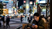 Young man of East Asian descent wearing casual dark clothing, sitting outdoors on a bench in an urban nighttime setting, focused on his smartphone. Busy city street with blurred pedestrians and illuminated storefronts in background. Concept for mobile technology, urban lifestyle, connectivity, and evening commute.