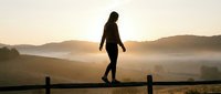 Young woman in athletic wear standing on wooden railing at sunrise, overlooking misty valley landscape. Silhouette figure for wellness, fitness, and outdoor lifestyle concepts.