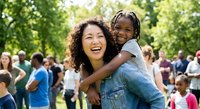 Happy mixed-race mother giving piggyback ride to smiling African American daughter at outdoor community event with diverse families in sunny park.