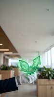 Businessman in formal attire holding green leaf and upward arrow symbol in modern office environment with plants, representing sustainable business growth and eco-friendly corporate strategy.
