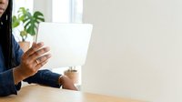 Young Black woman in professional business attire working on laptop at wooden desk with potted plant, bright office environment with natural light.