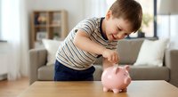 Young boy in striped shirt putting coin into pink piggy bank at home, learning financial literacy and saving money habits in modern living room.