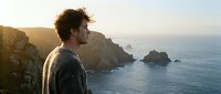 Young man with curly hair gazing at ocean sunset from dramatic coastal cliffs, peaceful moment of contemplation and connection with nature.