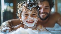 Joyful young boy laughing during bath time with soap bubbles in hair and on face, showing pure childhood happiness while parent watches with smile in background.
