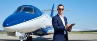 Professional businessman in suit with tablet standing confidently next to luxury private jet on airport tarmac under clear blue sky for corporate travel.