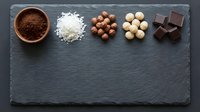 Chocolate making ingredients arranged on dark slate board including cocoa powder, coconut flakes, hazelnuts, white chocolate chips and dark chocolate pieces.