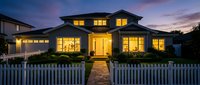 Luxury suburban home with warm interior lighting at twilight, featuring white picket fence and dramatic purple sky for real estate marketing.