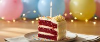 Red velvet cake slice with white frosting and single lit candle on plate with colorful balloons and fairy lights for birthday celebration party dessert.