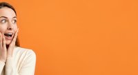 Excited young woman with long brown hair wearing cream sweater, touching face with both hands, expressing surprise and joy against vibrant orange background for advertising and marketing campaigns.