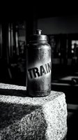 Metallic sports water bottle with train text label sitting on concrete surface in gym environment, black and white photography.