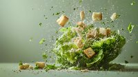 Fresh green lettuce leaves and crunchy croutons exploding in dynamic composition with water drops and particles against soft green background. Food photography.