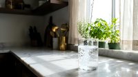 Clean glass of fresh water on marble kitchen counter near window with green plants creating healthy hydration lifestyle scene.