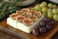 Fresh figs arranged on soft brie cheese with purple and green grapes on wooden cutting board, garnished with herbs for gourmet appetizer presentation.