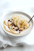 Creamy yogurt bowl topped with fresh banana slices, caramel drizzle and dark chocolate shavings served on white background, healthy breakfast concept.